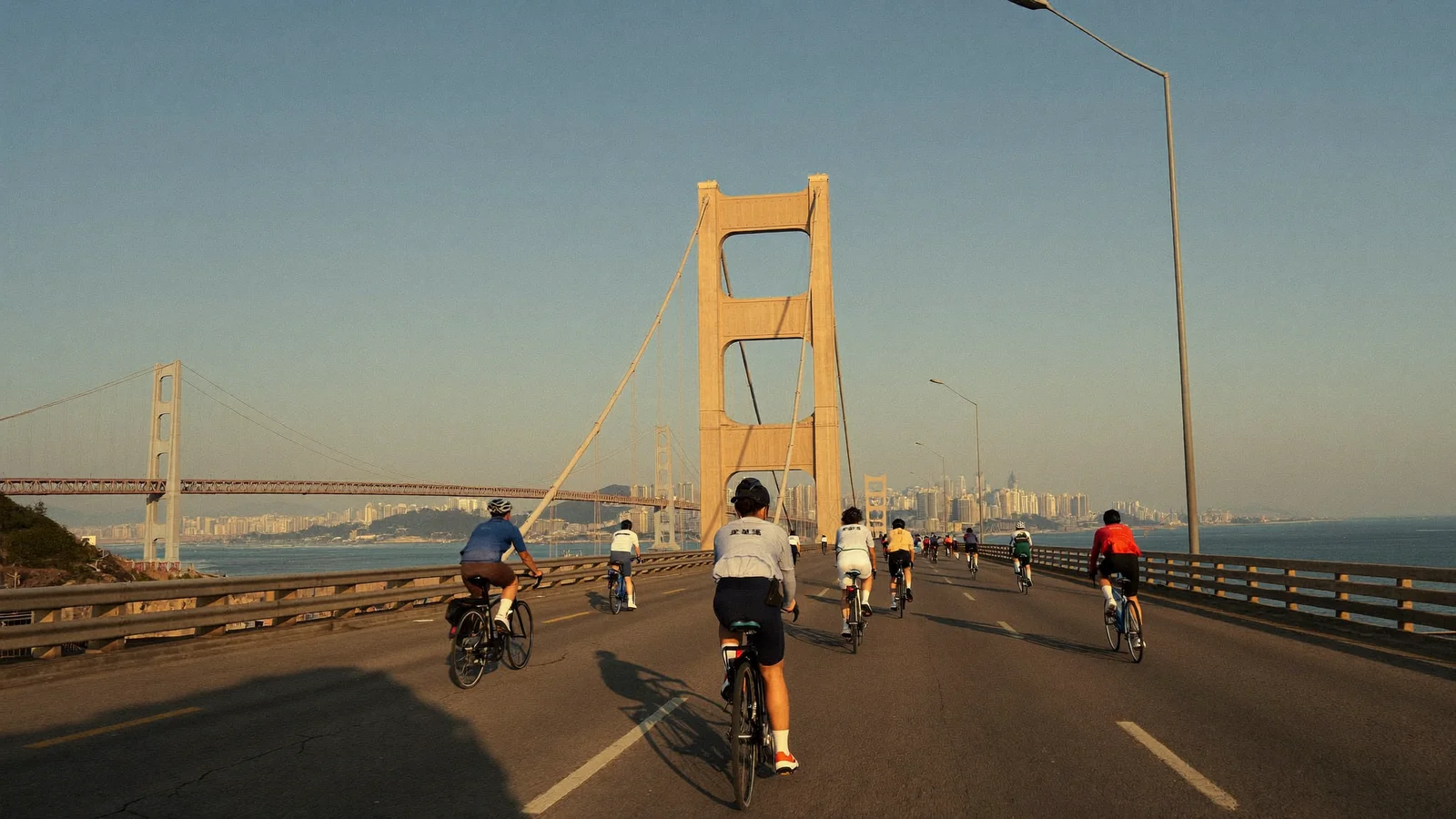 Illustrative image of cyclists in bright jerseys riding across a long suspension bridge over Hong Kong harbour on a clear day, not a photo of the actual event