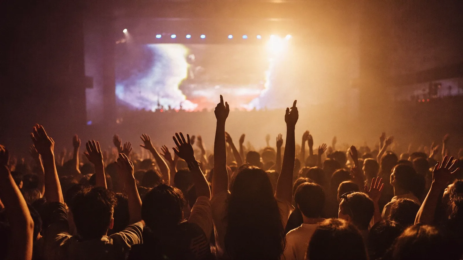 Silhouetted concert crowd with arms raised toward warm stage lights in a generic indoor hall