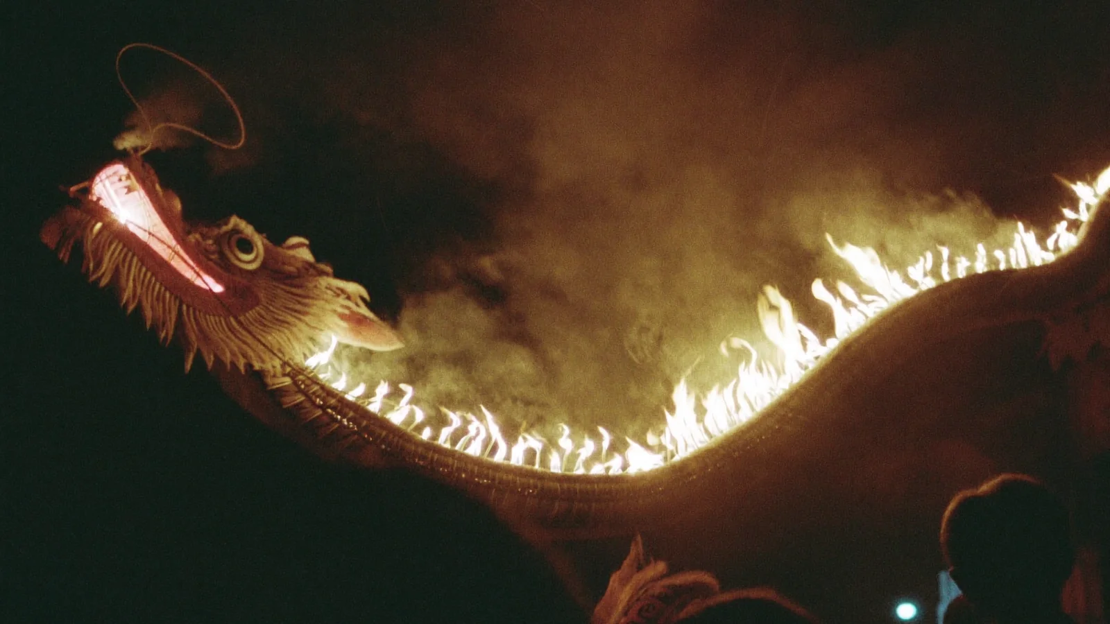 Illustrative image of a straw fire dragon studded with burning incense, glowing and trailing smoke against a night sky